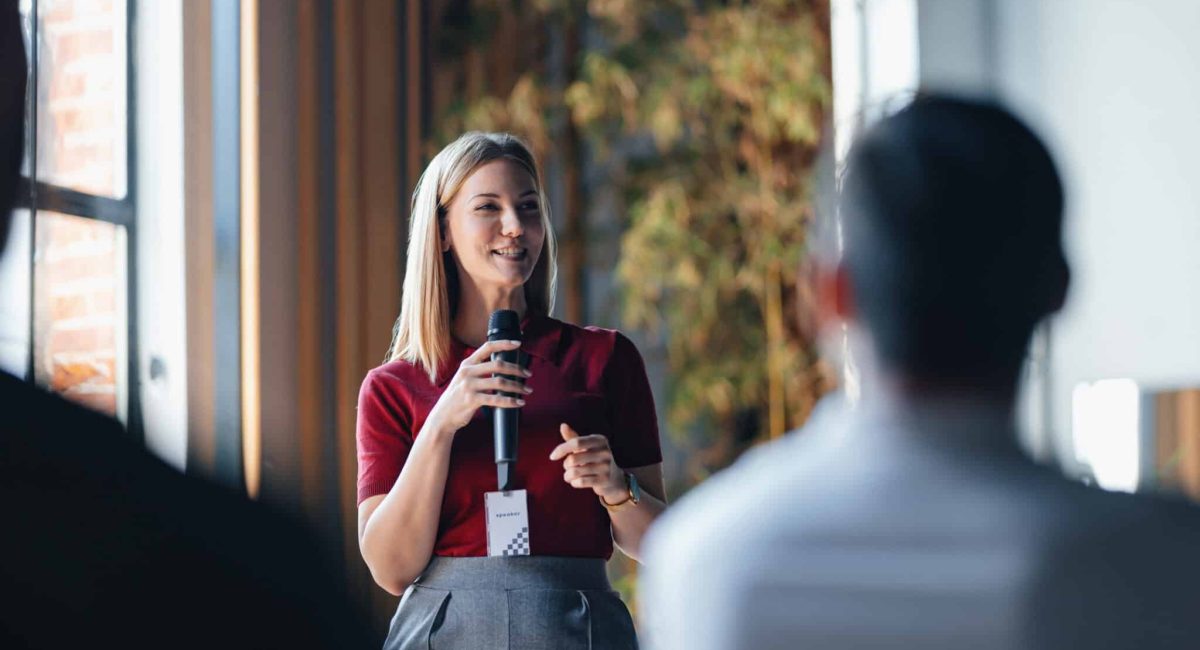 A confident woman delivers a speech to an attentive audience at a professional networking event. The setting suggests collaboration and knowledge sharing among business professionals.