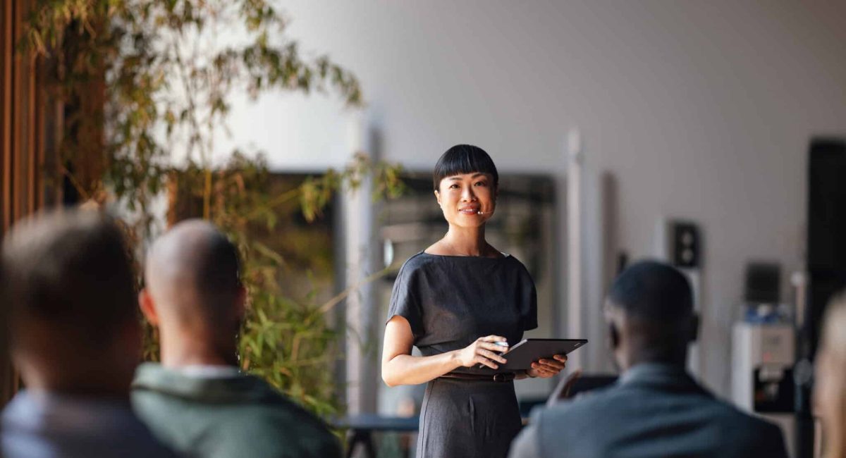 A confident presenter leads a business conference, engaging a diverse audience. The speaker is using a tablet and microphone, ensuring effective communication and interaction in a modern professional setting.